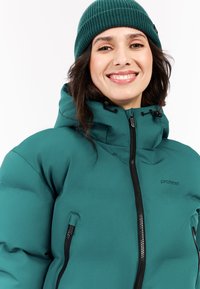 Teal pufferjack met een hoge kraag, ritssluiting en twee zijzakken; gecombineerd met een donkergroene gebreide beanie. Zwart logo-detail zichtbaar.