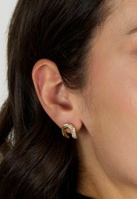 Orecchini color oro con un design curvo, impreziositi da cristalli trasparenti, che presentano una superficie texturizzata e una finitura lucida.