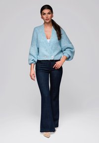 Ljusblå stickad kort tröja med V-hals,vida ärmar och två knappar i midjan, i kombination med mörka utsvängda jeans och beige skor.