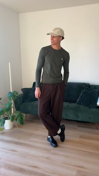 Camicia verde oliva a maniche lunghe, pantaloni marroni a gamba larga e scarpe nere, con un cappello beige. In piedi davanti a un divano verde e una pianta.