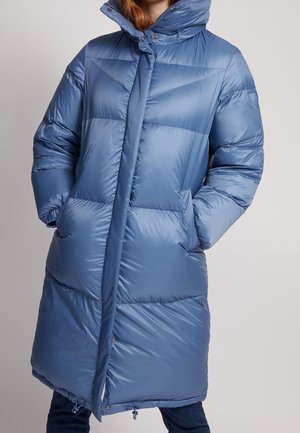 Femme portant un long manteau d'hiver rembourré bleu avec un col haut et les mains dans les poches, se tenant contre un fond uni.