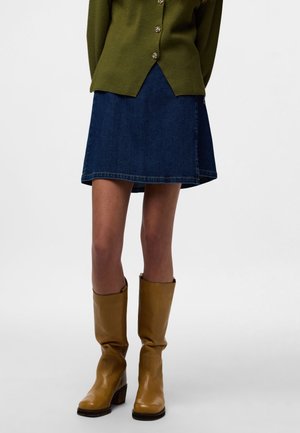 Kvinde iført en mørkeblå denimnederdel, olivengrøn knappet sweater og brune læderknæstøvler mod en hvid baggrund.