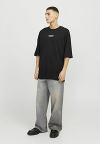 Czarna, oversize t-shirt z grafiką "ICONIC" na klatce piersiowej, zestawiona z jasnoszarymi spodniami o szerokich nogawkach i czarnymi, masywnymi butami. Gładka tekstura materiału.