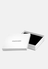 Witte rechthoekige doos met een zwarte binnenkant, voorzien van het "CALVIN KLEIN" logo op de deksel in zwarte letters. Geopend, met een zachte zwarte voering.