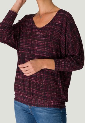 Frau trägt ein langärmliges Oberteil mit V-Ausschnitt in Burgunderrot und Schwarz mit Muster, dazu blaue Jeans, eine Hand nahe am Gesicht erhoben und die andere Hand in der Taille ruhend.