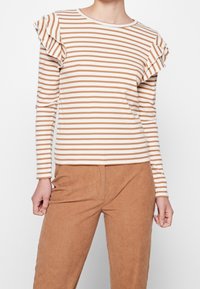 Gestreifter Langarm-Oberteil in Creme und Braun mit Rüschenakzenten an den Schultern. Kombiniert mit hellbraunen Cordhosen, die eine gerippte Textur aufweisen.