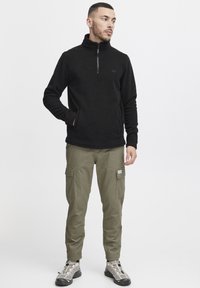 Suéter de forro polar negro con cuello de media cremallera, combinado con pantalones cargo verde oliva que cuentan con bolsillos laterales y un parche. El calzado son zapatillas grises.