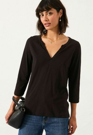 Schwarzes Langarmshirt mit V-Ausschnitt und Knopfdetails. Weicher Stoff, lockere Passform, kombiniert mit blauen Jeans und einer kleinen schwarzen Handtasche.