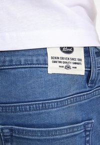 Modré denimové džíny s texturačním povrchem, s bílou etiketou na pasu, která zobrazuje název značky a pokyny k údržbě.