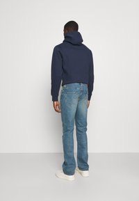 Mężczyzna odwrócony plecami, ubrany w granatowy kaptur, jasnoniebieskie dżinsy i białe trampki, stoi na jednolitym białym tle.