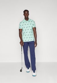 Light blue polo shirt with golf club pattern, paired with navy trousers and white golf shoes. Standing with a golf club in hand.
