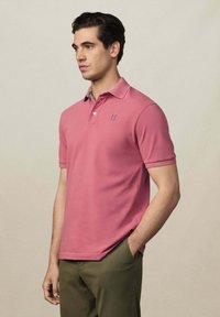 Ružová polo košeľa z bavlny s krátkymi rukávmi, na hrudi s modrým logom a s vzorovaným golierom. Nosená s zelenými nohavicami.