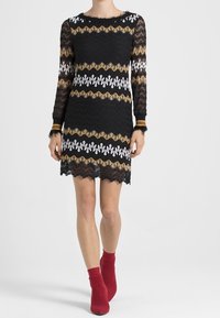 Svart spetsklänning med långa ärmar, prydd med vågiga mönster i vitt och guld. Fållkanten är vågig. Kombinerad med röda ankelskor.