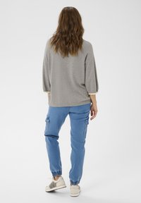 Ženska z dolgimi valovitimi lasmi, oblečena v sivo pulover, modre cargo hlače in bele superge, stoji pravokotno ob beli ozadju.