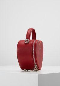 Rode leren tas in de vorm van een hart met een handvat aan de bovenkant, zilveren rits en metalen voetjes op een witte ondergrond. Bevat logo aan één kant.