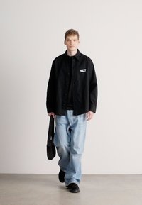 Μαύρο πουκάμισο με κουμπιά πάνω από μαύρο μπλουζάκι, ανοιχτό μπλε άνετο τζιν, μαύρα παπούτσια και μαύρη τσάντα ώμου. Ομαλές υφές.