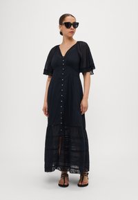Robe maxi noire avec un devant à boutons, des manches courtes évasées, une bordure en dentelle et une jupe à volants ; associée à des lunettes de soleil noires et des sandales.