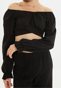 Schwarzes, schulterfreies Oberteil mit langen Ärmeln, gerafften Bündchen und einem gedrehten Vorderdesign, aus glattem Stoff mit matter Oberfläche.
