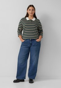 Frau steht mit den Händen in den Taschen, trägt ein gestreiftes Langarmpolohemd, weite blaue Jeans und schwarze Schuhe vor einem einfarbigen Hintergrund.