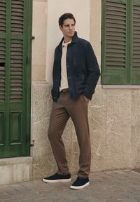 Marineblauwe jas over een crèmekleurig shirt, bruine gestreepte broek en donkere sneakers. De achtergrond toont een groen raamluik en een gestructureerde muur.