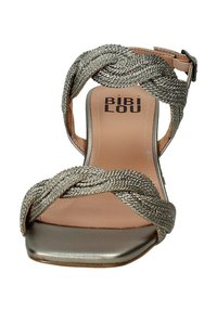 Sandalias trenzadas plateadas con puntera cuadrada y una correa metálica. La plantilla es de color beige con un logotipo negro en la suela. Correa ajustable en el tobillo.