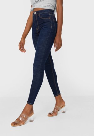 Femme portant un jean skinny taille haute bleu foncé et des sandales à talons avec bride transparente, debout devant un fond clair uni.