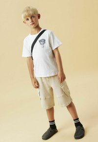 T-shirt blanche avec imprimé noir, shorts cargo beige, chaussures noires et chaussettes rayées. Fond neutre, l'enfant se tient dans une pose décontractée.
