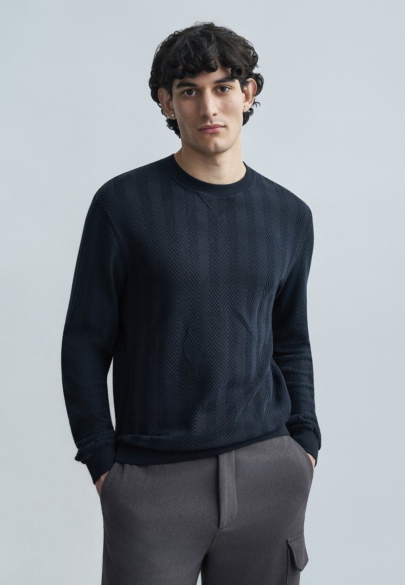 Μαύρο πλεκτό πουλόβερ με σχέδιο ψαροκόκκαλο, λαι μόκοψη στρογγυλή και ριμπ μανσέτες, σε συνδυασμό με γκρι παντελόνι. Λεία υφή και εφαρμοστή γραμμή.