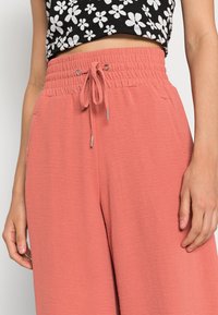 Persikofärgade vida byxor med elastisk midja och dragsko. Mjuk tygtextur med sidfickor; modellen bär en blommig kort topp.