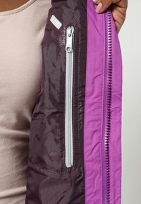 Interior de chaqueta aislante púrpura y marrón oscuro con un bolsillo vertical con cremallera en gris claro, con textura suave y costuras distintivas.