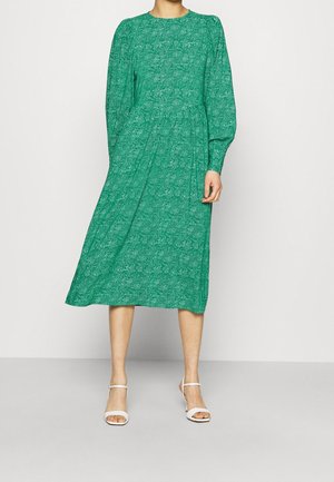 Femme portant une robe midi verte à manches longues avec des motifs subtils et des talons blancs ouverts, debout devant un fond clair uni.