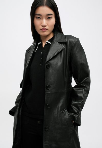 Cappotto in pelle nera con colletto a punte, bottoni frontali e tasche laterali, indossato sopra un maglione nero con colletto rifinito in bianco.