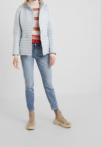 Světle modrá zateplená bunda s horizontálním prošíváním, zkombinovaná se pruhovaným svetrem, modrými džíny a hnědými turistickými botami s tkaničkami.