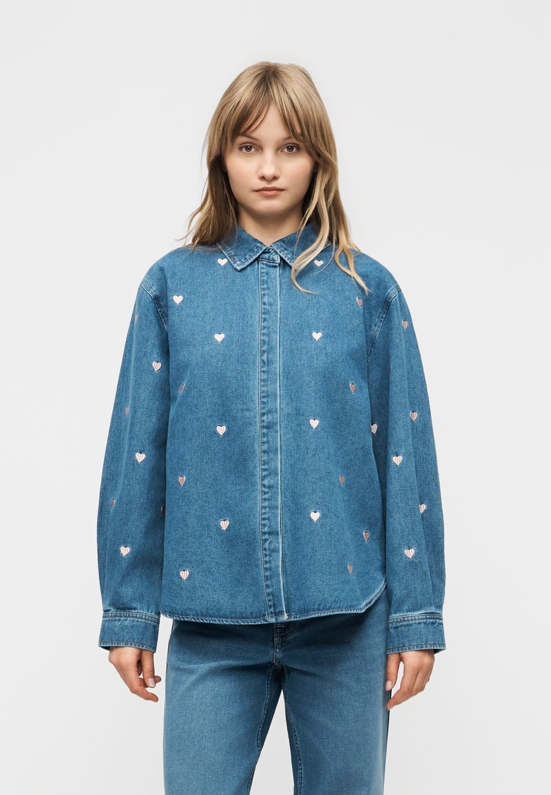 Mujer joven vistiendo una camisa de mezclilla azul con pequeños patrones de corazones rosas y jeans de mezclilla a juego, de pie frente a un fondo liso.