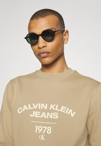 Occhiali da sole neri con un design a montatura spessa e rotonda. Indossati sopra una felpa beige con testo bianco: "CALVIN KLEIN JEANS 1978."