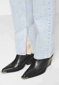 Botines de cuero negro con punteras puntiagudas, detalles de tachuelas plateadas y tacón grueso, combinados con jeans azul claro que tienen cremalleras en el dobladillo.