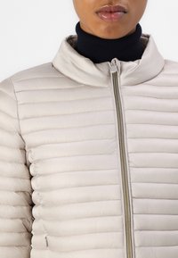 Lys beige pufferjakke med lynlås, stående krave og quiltet tekstur. Har horisontal syning og en glat overflade.