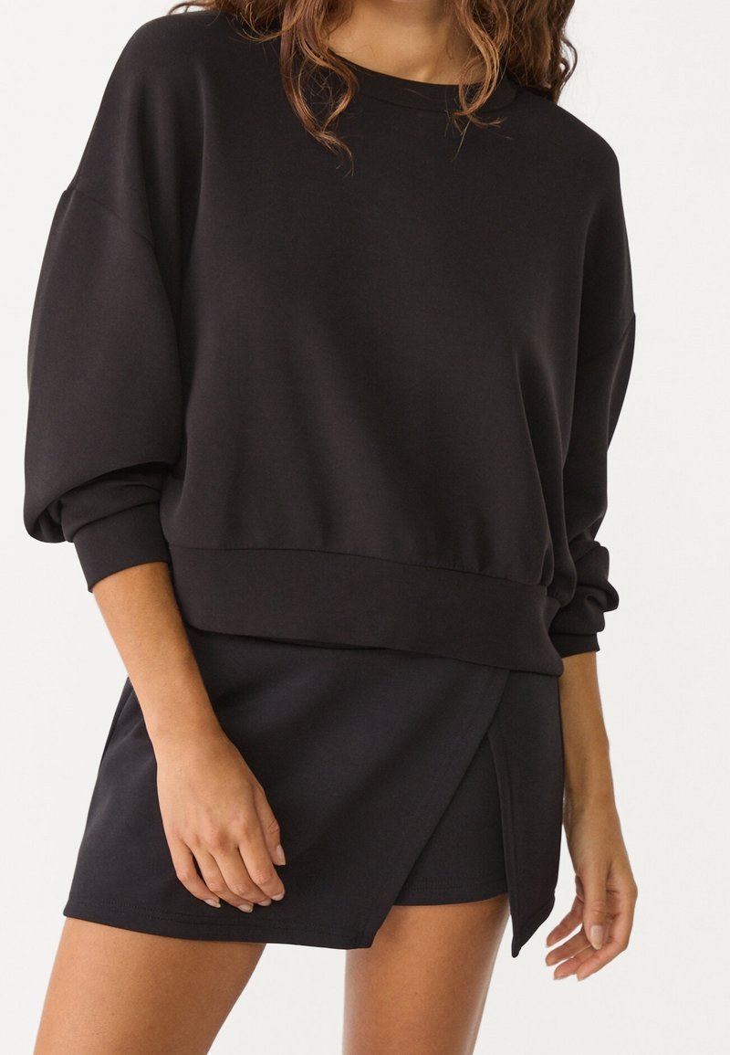 Sudadera negra de manga larga con hombros caídos y puños acanalados, combinada con una falda envolvente negra a juego que presenta un dobladillo en ángulo.