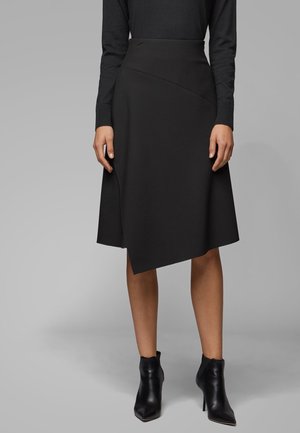 Nainen, joka on pukeutunut mustaan epäsymmetriseen polvipituiseen hameeseen, mustaan pitkähihaiseen puseroon ja mustiin nilkkureihin, seisoo harmaan taustan edessä.