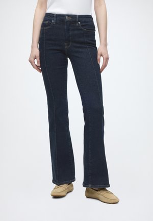 Mørkeblå jeans med lige ben og forlommer, båret med beige loafers og en hvid top på en person, der står.