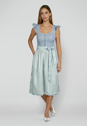 Frau trägt ein ärmelloses hellblaues Kleid mit gerafften Schultern, einem Reißverschluss vorne, einem gebundenen Gürtel in der Taille und weißen, spitz zulaufenden High Heels.