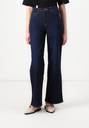 Femme portant un jean flare taille haute bleu foncé avec des coutures contrastantes, associé à des chaussures plates beiges à bout pointu avec des brides à boucle.