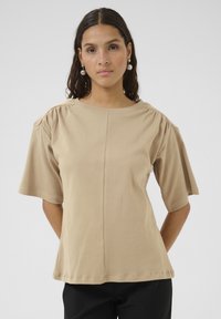 Beige T-shirt met korte mouwen en een ronde halslijn, met geplooide schouderdetails en een losse pasvorm. Gemaakt van zachte, lichte stof.