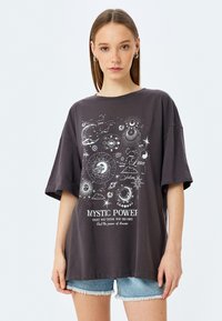 T-shirt oversized gris foncé à manches courtes avec des graphiques célestes blancs et le texte "MYSTIC POWER" imprimé sur le devant.