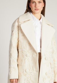 Abrigo de lana beige con intrincados bordados florales y adornos. Amplios solapas, cierre de doble botonadura y tejido texturizado.