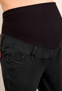 Čierne tehotenské nohavice s elastickým čiernym pásom. Obsahujú bočné vrecká a majú priliehavý strih. Materiál sa zdá byť mäkký a odolný.