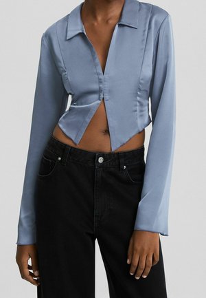 Helles blaues Satin-Croppshirt mit langen Ärmeln, einem spitzen Kragen und einem tiefen V-Ausschnitt, kombiniert mit hochtaillierten schwarzen Jeans.