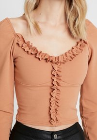 Hnědý cropped top s dlouhými rukávy, s volánkovým výstřihem a předním detailem. Vyroben z hladké látky s přiléhavým střihem.