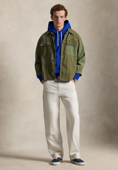 Jeune homme portant une veste patchwork verte sur un sweat à capuche bleu, un pantalon blanc et des baskets bleues, debout devant un fond beige uni.