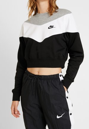 Personne portant un sweat-shirt court Nike noir, blanc et gris et un pantalon Nike noir avec des boutons-pression blancs sur les côtés.